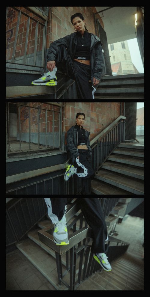 Woman in black jacket and pants poses on brick stairs, wearing grey and neon green sneakers outdoors, urban setting, overcast day.