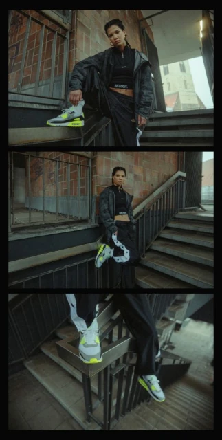 Woman in black jacket and pants poses on brick stairs, wearing grey and neon green sneakers outdoors, urban setting, overcast day.