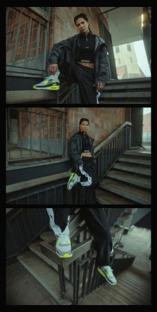 Woman in black jacket and pants poses on brick stairs, wearing grey and neon green sneakers outdoors, urban setting, overcast day.