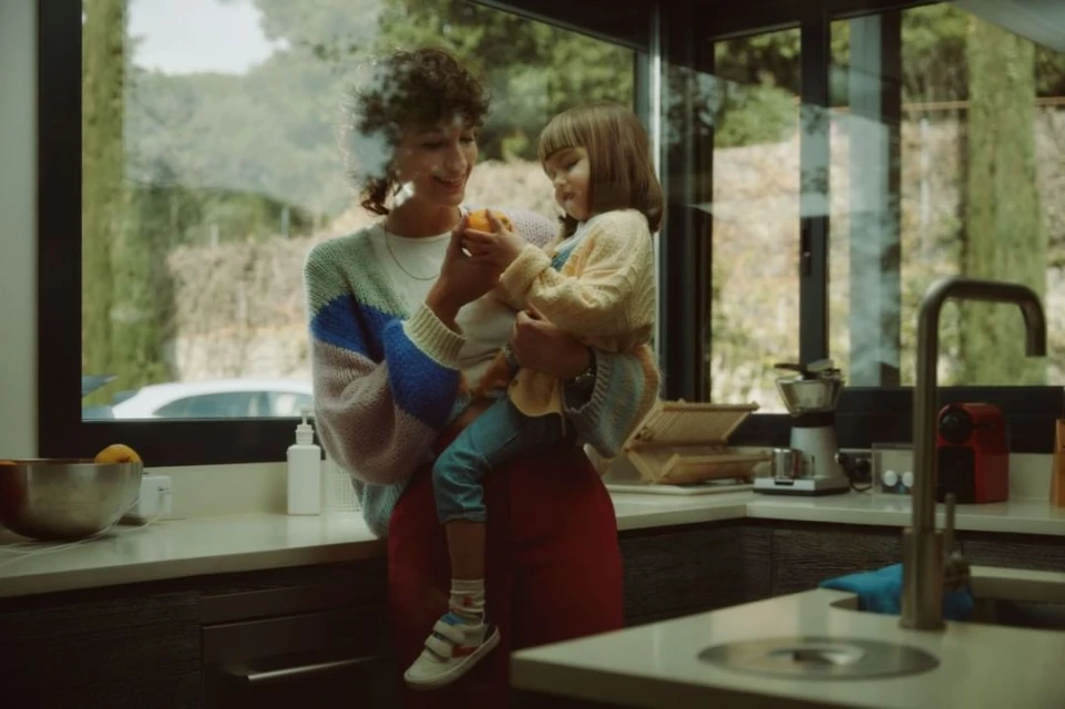 Mother and child indoors holding an orange, near a kitchen counter. Light, airy scene with natural light through the window. Casual lifestyle.