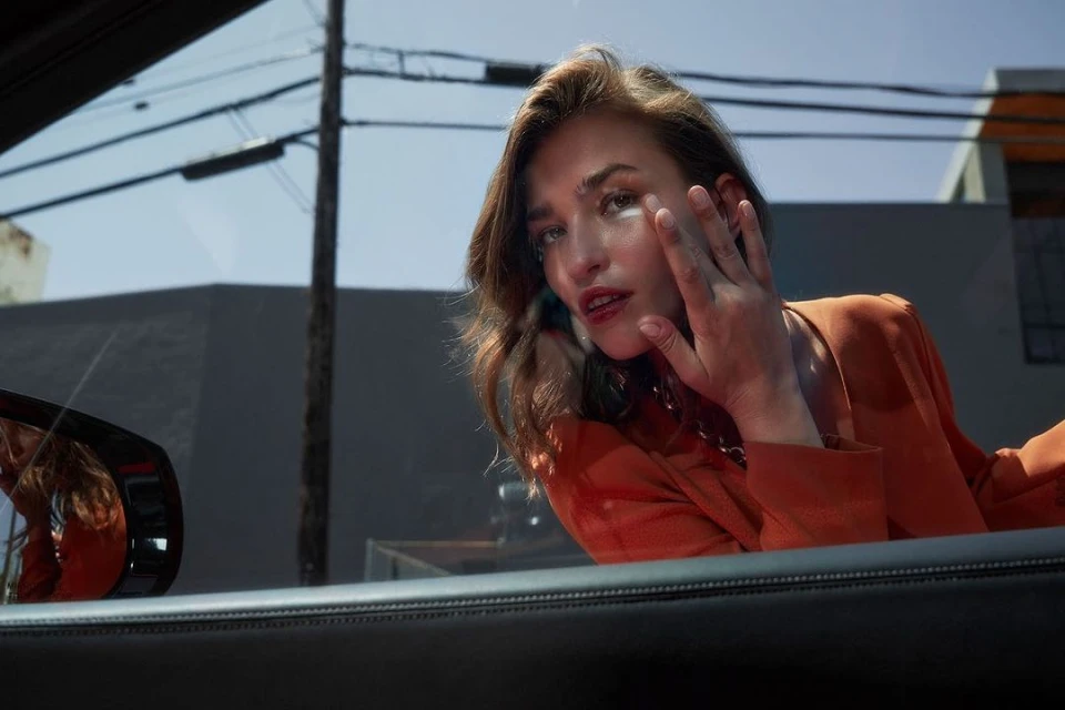 Portrait of a young woman with hand near face, shot inside a car on sunny day with buildings and telephone poles in the background.