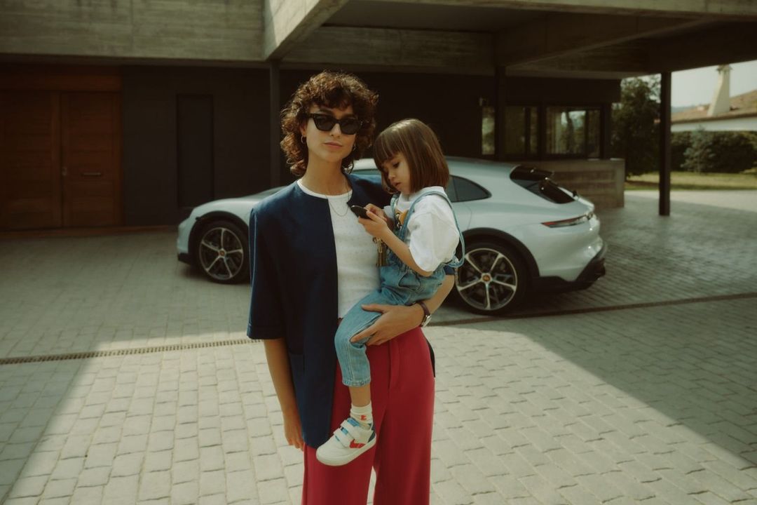 Woman in a color-blocked blazer holds a child in overalls, both looking at a mobile phone. An SUV is visible in the background.