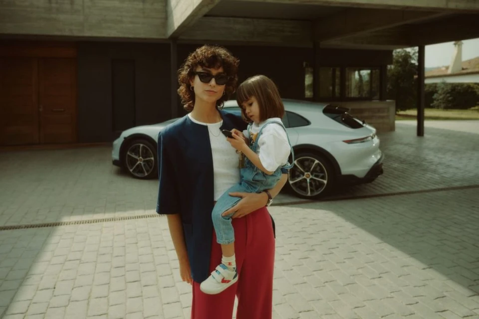 Woman in a color-blocked blazer holds a child in overalls, both looking at a mobile phone. An SUV is visible in the background.