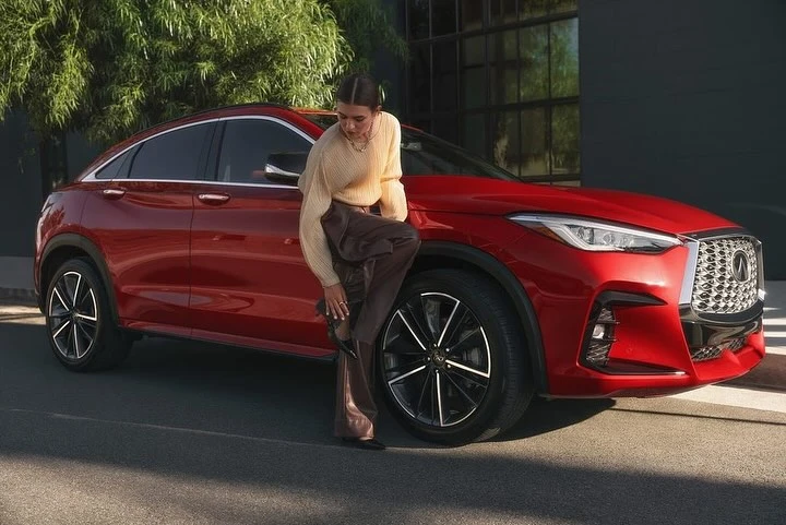 A red Infiniti QX55 coupe SUV is showcased with a woman in neutral tones leaning casually against its hood outdoors, a stylish automotive scene.