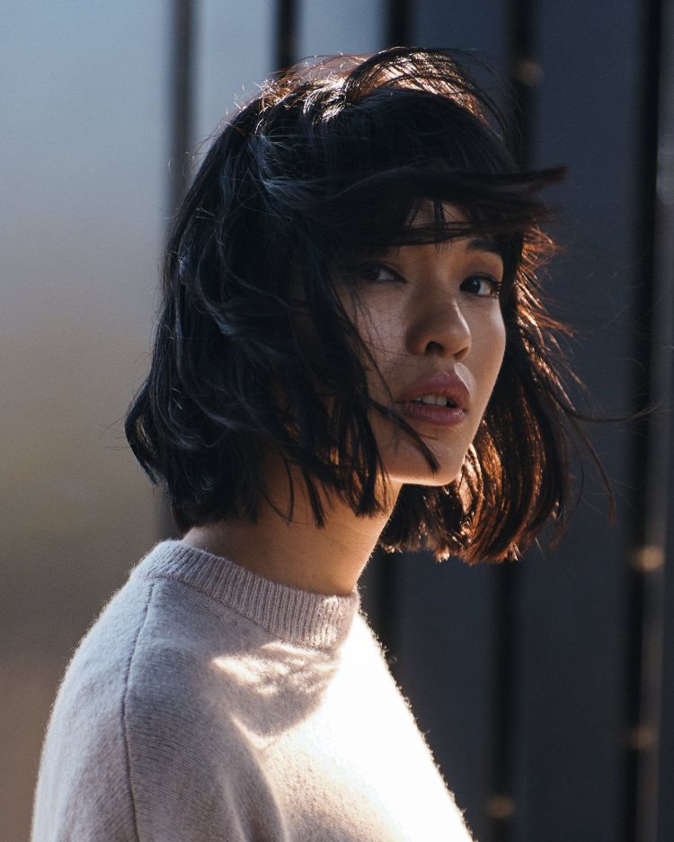 Close-up shot of an adult woman with short dark hair and light skin wearing a sweater, posing in front of a building outdoors.