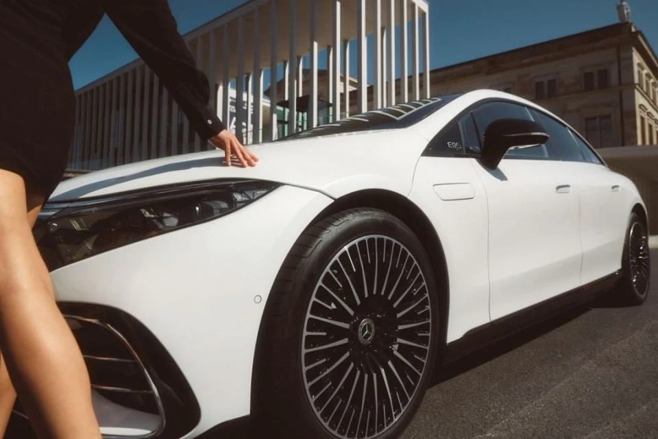 White electric sedan parked outdoors with an adult woman's hand resting on the hood, set against a modern building and blue sky.