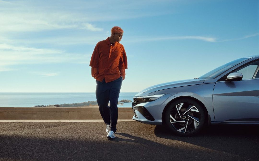 A stylish adult male walks near a parked silver car with the ocean and sky visible in the background on a sunny day.