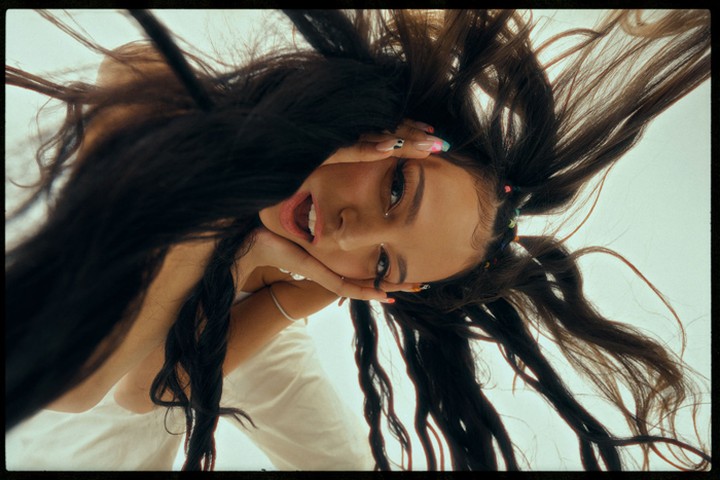 Low-angle shot of a young woman with long, dark hair framing her face, posing with her hands and displaying painted nails.