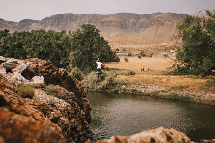 A person jumps into a river from a rocky cliff, with trees and mountains in the background under a bright sky. Adventure and nature.