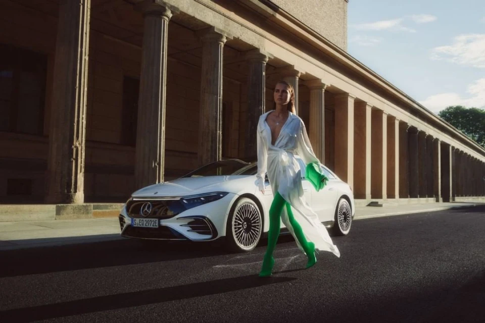Fashion shot of a woman in white dress with car. Woman walks on asphalt street in front of white electric car and stone building with columns.