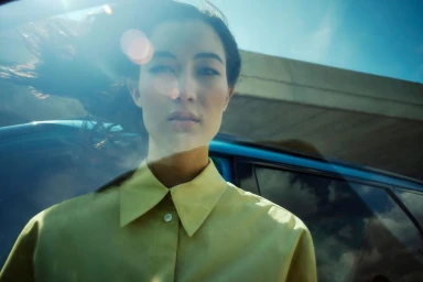 Woman wearing a yellow button-down shirt standing in front of a blue car under a clear blue sky captured by David Daub