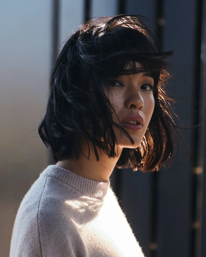 Portrait of a woman with dark short hair and bangs wearing a cream sweater against a dark slatted wooden background