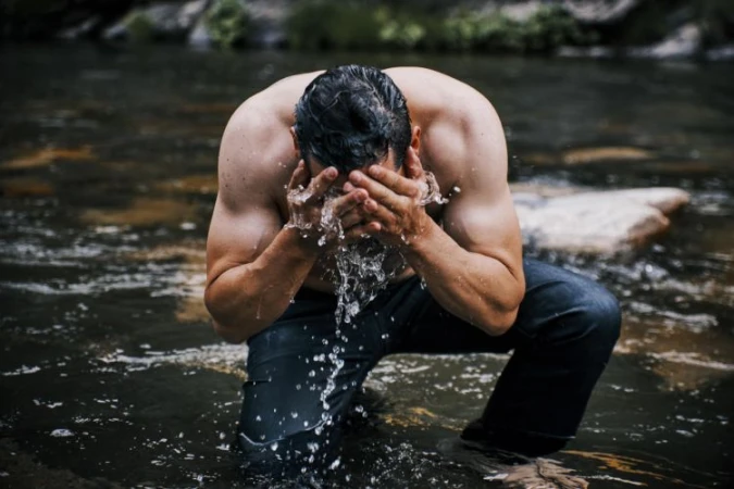 Shirtless man kneeling in a river splashing water onto his face