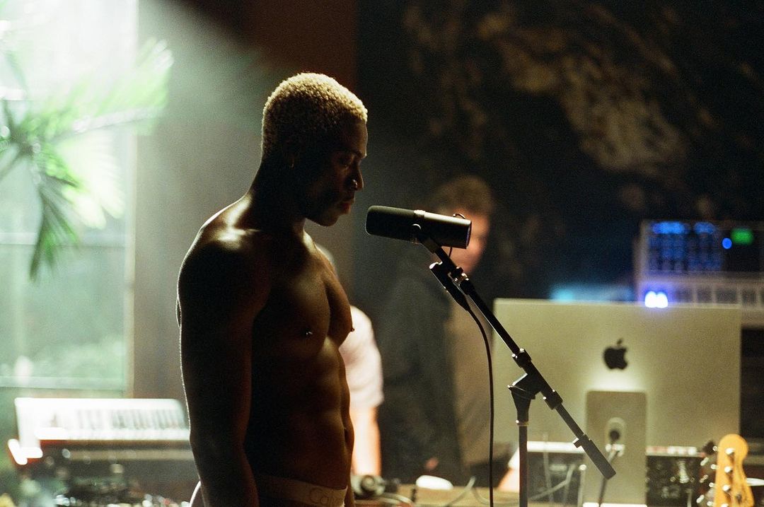 Close-up of an adult man singing into a microphone in a dimly lit music studio. Keyboard, computer screen and other equipment visible.