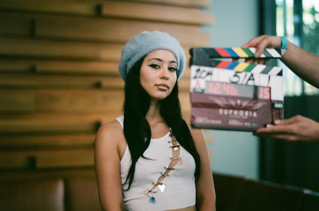 Female model with a clapperboard, advertising the TV series Euphoria. She is wearing a beret, a white top and statement makeup.