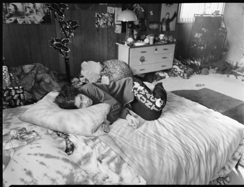 Teenage girl with long hair lounges on a bed in a black and white medium shot. Posters and cluttered shelves adorn the wood-paneled walls.
