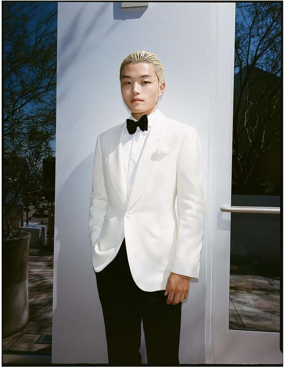 Portrait of a young man in a white suit with a black bow tie standing in front of a wall with an outdoor view.