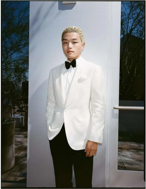 Portrait of a young man in a white suit with a black bow tie standing in front of a wall with an outdoor view.