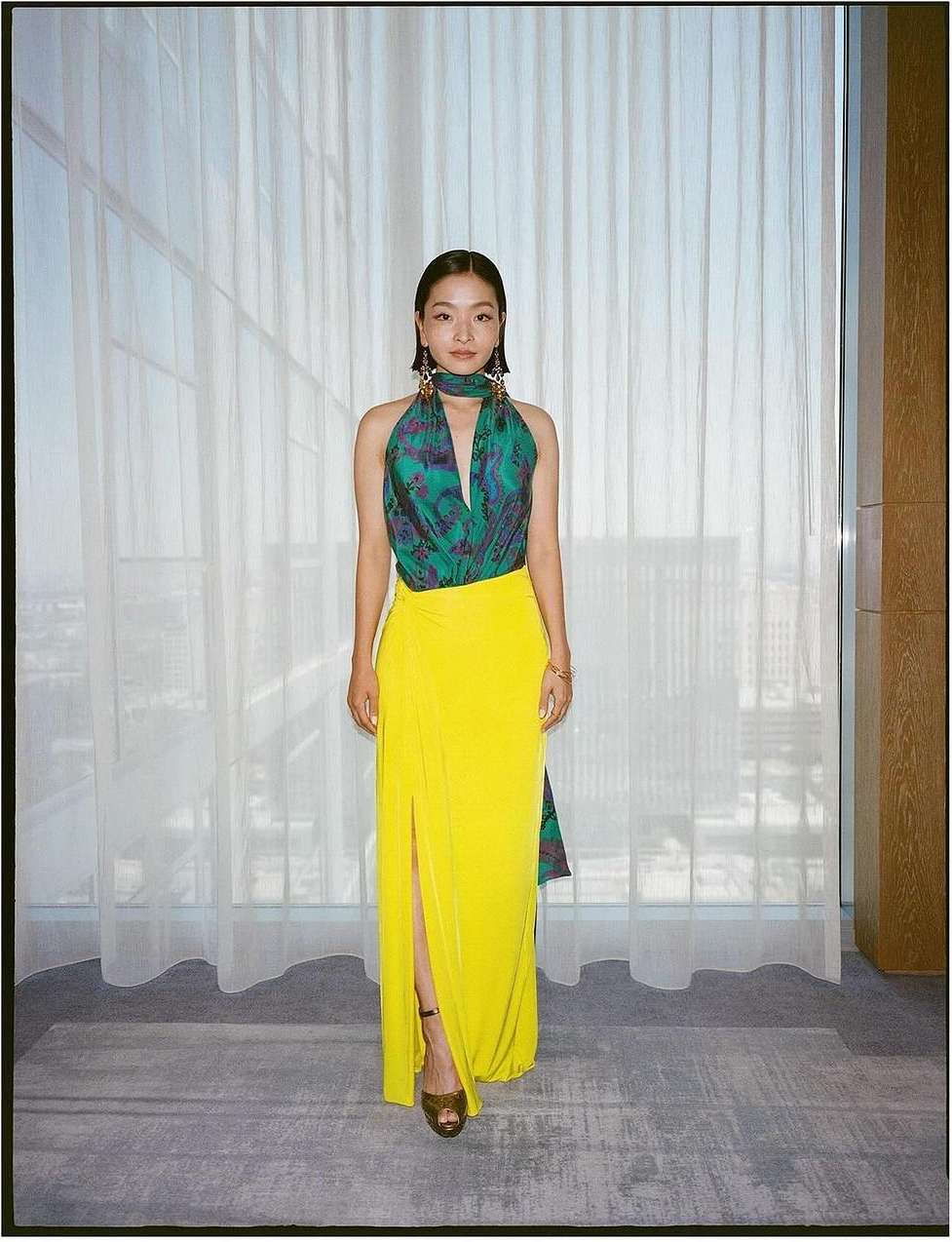 Elegant Asian woman in a halter top and long yellow skirt poses in front of a window with sheer white curtains; fashion portrait.