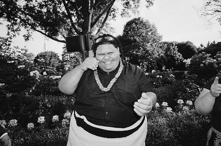 Black and white outdoor portrait of a smiling adult male wearing a floral lei and sunglasses with trees and a monument in the background.