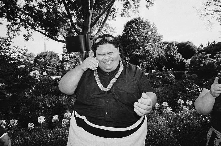 Black and white outdoor portrait of a smiling adult male wearing a floral lei and sunglasses with trees and a monument in the background.