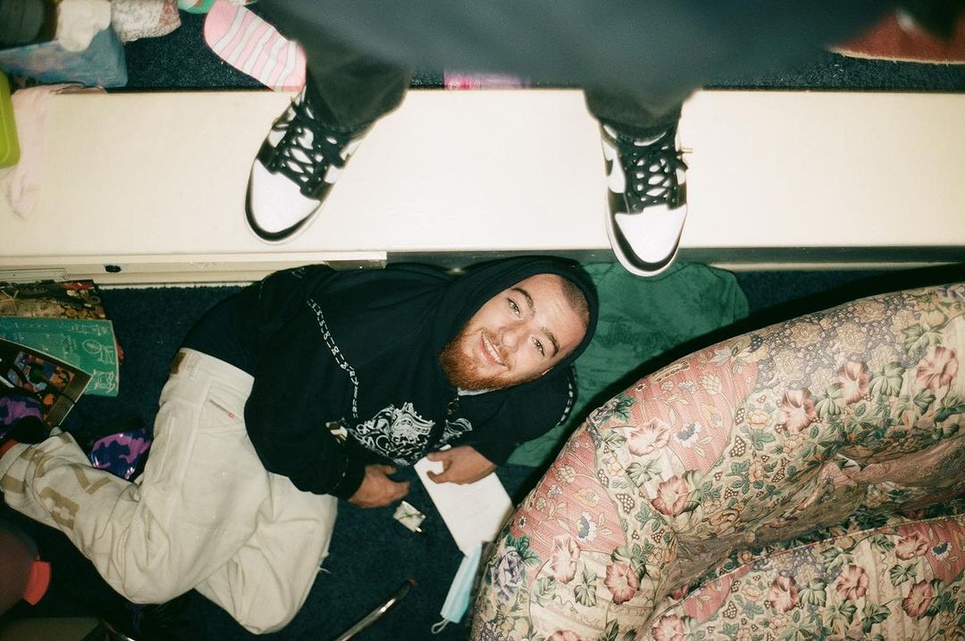 Overhead shot of smiling man in hoodie laying on floor, with another person's legs and sneakers visible near the top.