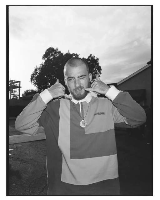 Black and white portrait of a man with a buzzcut and beard, wearing a long-sleeved shirt with patchwork design, outdoors with trees.