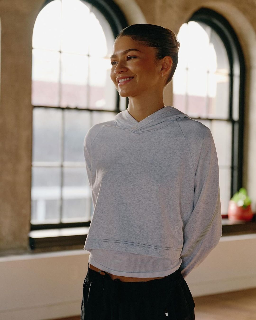 Zendaya poses near a window wearing a gray hooded sweatshirt and black pants, smiling softly with a serene expression.