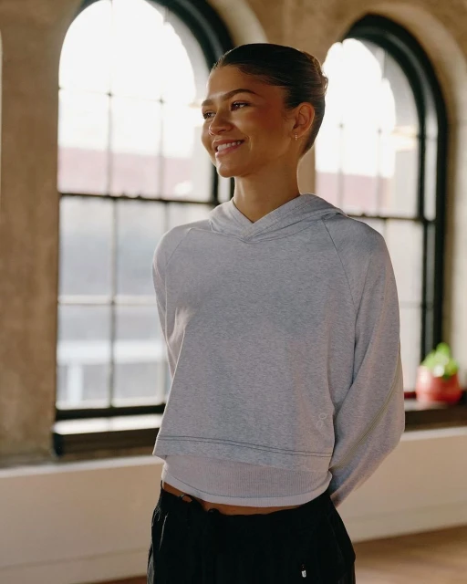 Zendaya poses near a window wearing a gray hooded sweatshirt and black pants, smiling softly with a serene expression.