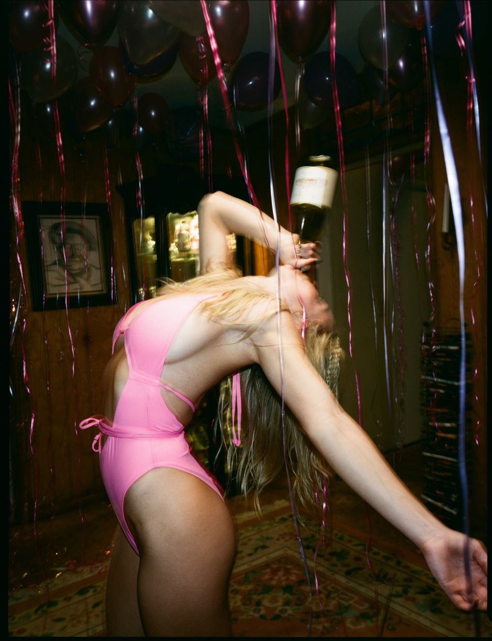 Blonde woman in pink swimsuit balances wine bottle on head surrounded by pink balloons and streamers in dimly lit room with wood paneling.