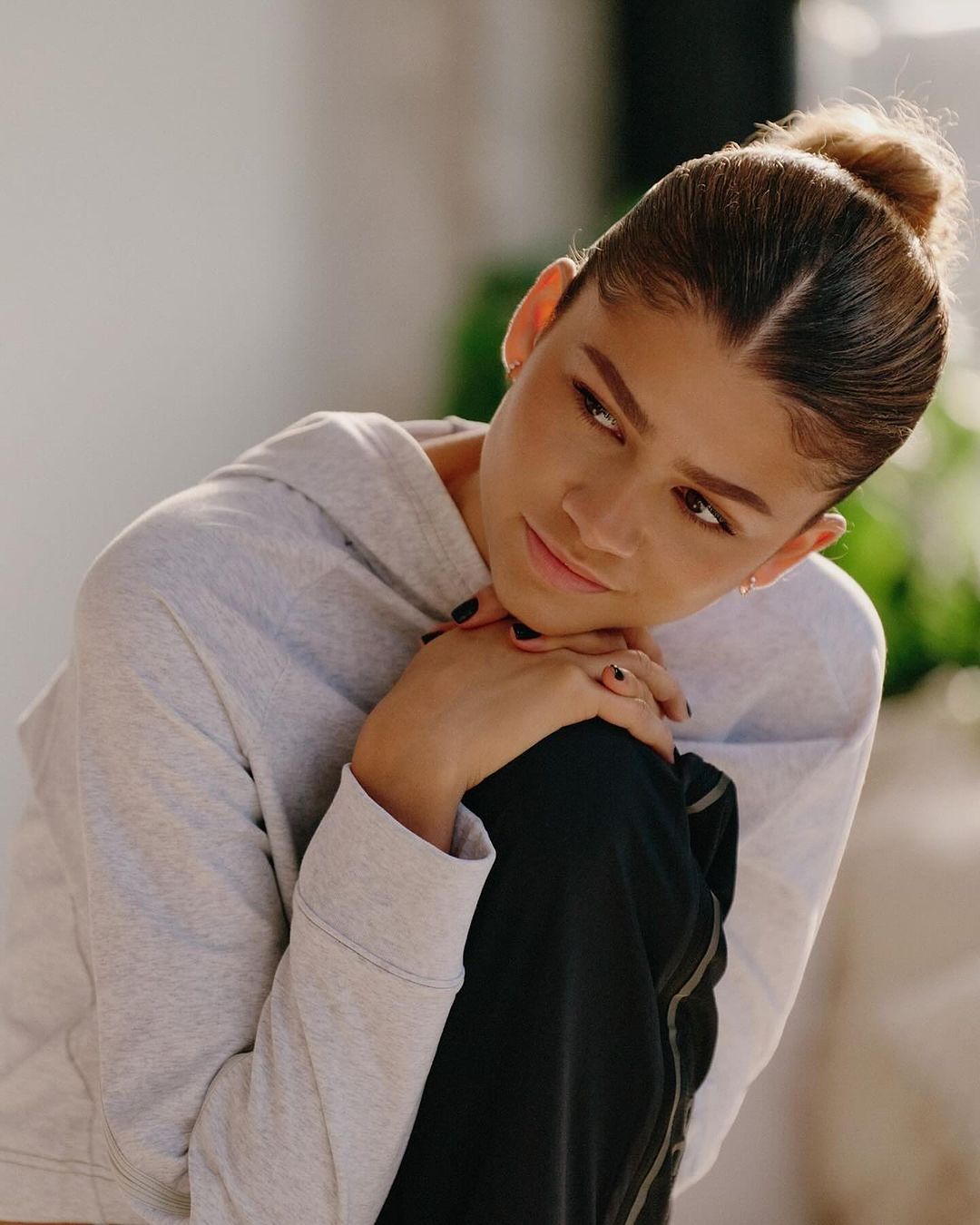 Close up portrait of Zendaya wearing a light grey hoodie with her hands clasped under her chin against a soft indoor background