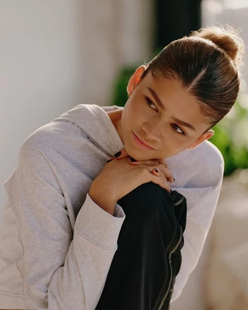Close up portrait of Zendaya wearing a light grey hoodie with her hands clasped under her chin against a soft indoor background