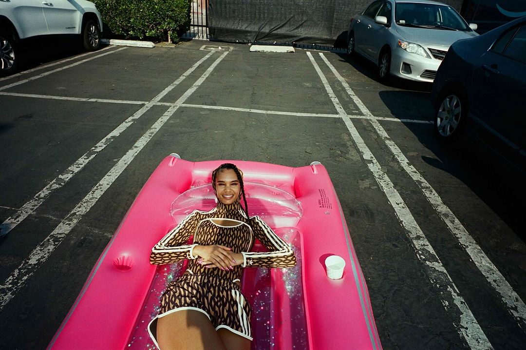 A woman in a patterned outfit poses on a pink inflatable in a parking lot, smiling and looking directly at the camera in outdoor lighting.