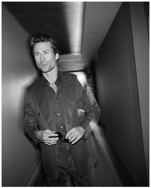 Black-and-white portrait of a stylish adult man in a suit, holding sunglasses in an indoor corridor with soft, blurred motion effect.