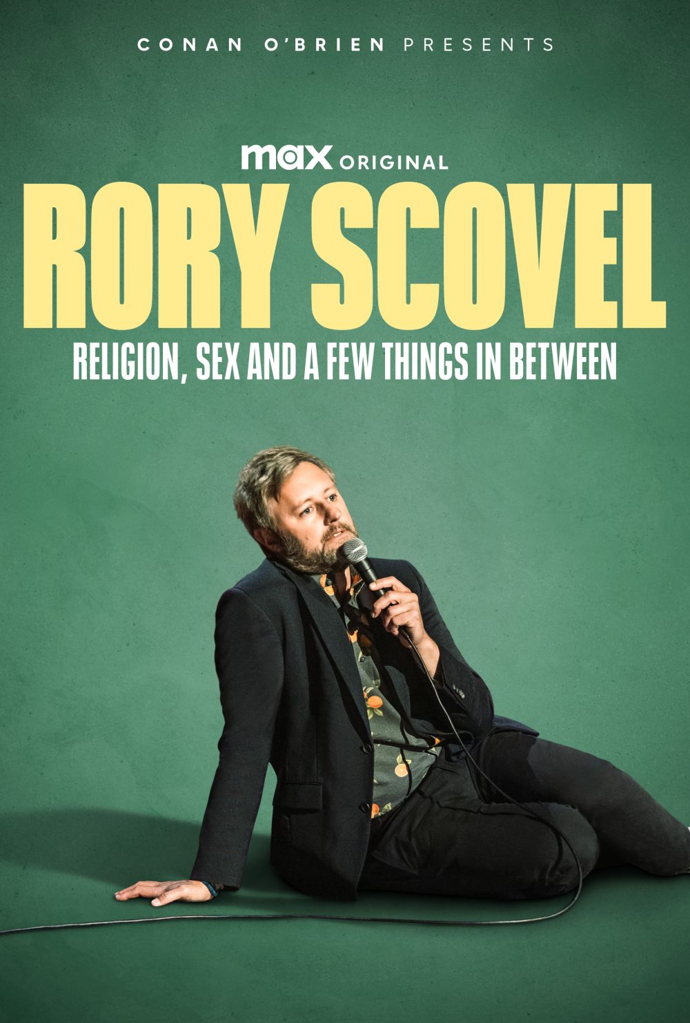 Rory Scovel, an adult man wearing a black blazer, sits against a green backdrop holding a microphone in a promotional studio shot.
