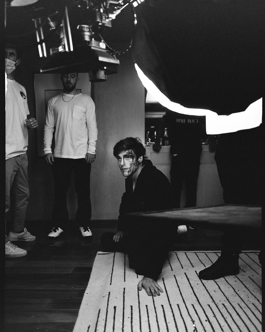Black and white image of a male model with makeup in a dark indoor setting, surrounded by crew members and film equipment on set.