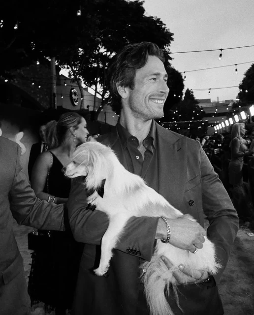 Black and white portrait of a smiling man holding a dog at an outdoor event, with other people visible in the background.