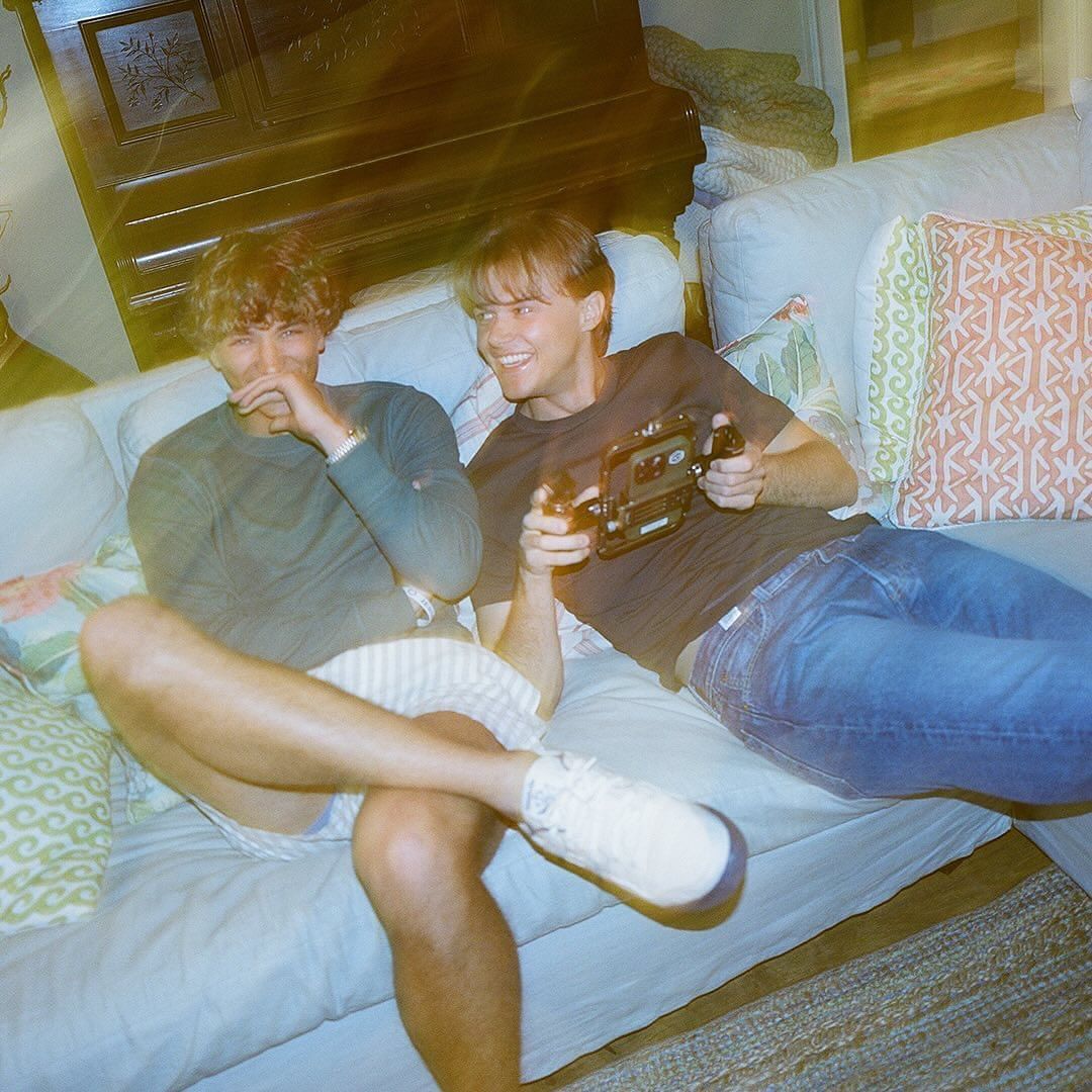 Two smiling young men seated together on a light blue couch, one holding a handheld camera, indoor, casual setting, warm lighting.