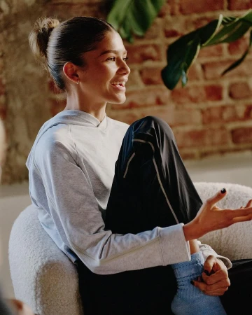Zendaya seated in a white armchair wearing a light grey hoodie and black trousers, captured by photographer Eddy Chen.