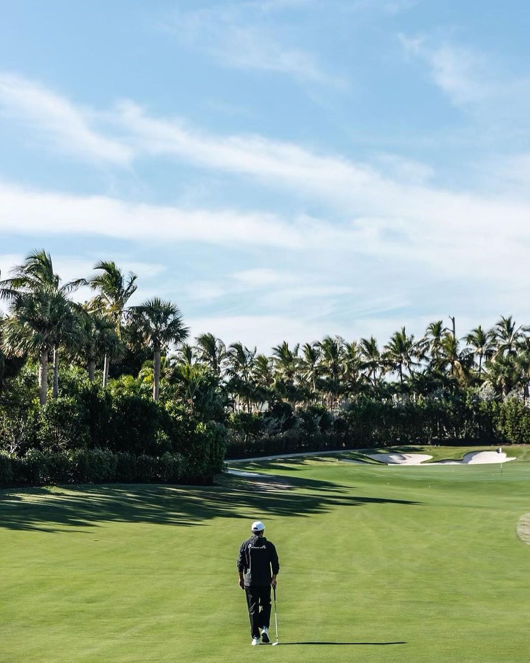 Person walking on golf course with trees and cloudy blue sky in the background. Golf club is carried in hand.