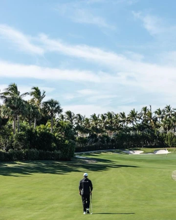 Person walking on golf course with trees and cloudy blue sky in the background. Golf club is carried in hand.