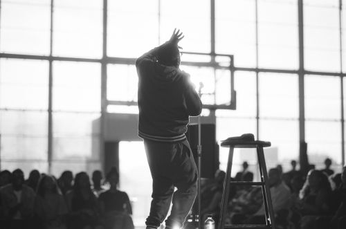 Monochrome photo of a person on stage, silhouetted against a bright window, addressing a seated audience with a stool nearby.