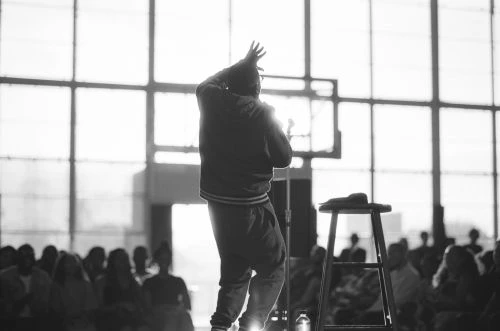 Monochrome photo of a person on stage, silhouetted against a bright window, addressing a seated audience with a stool nearby.