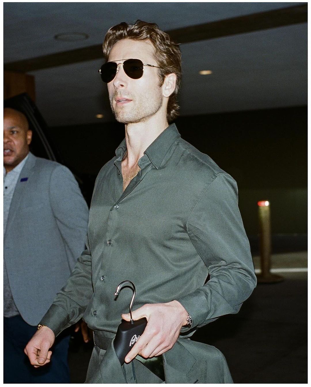 Man in dark green long-sleeve shirt and aviator sunglasses walking indoors with another person in background