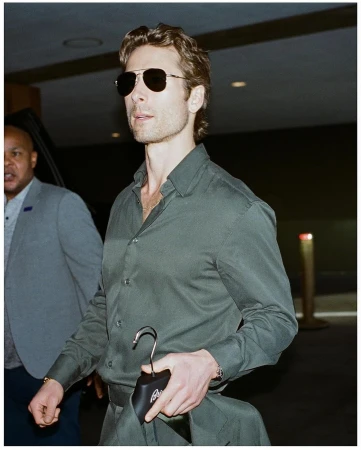 Man in dark green long-sleeve shirt and aviator sunglasses walking indoors with another person in background