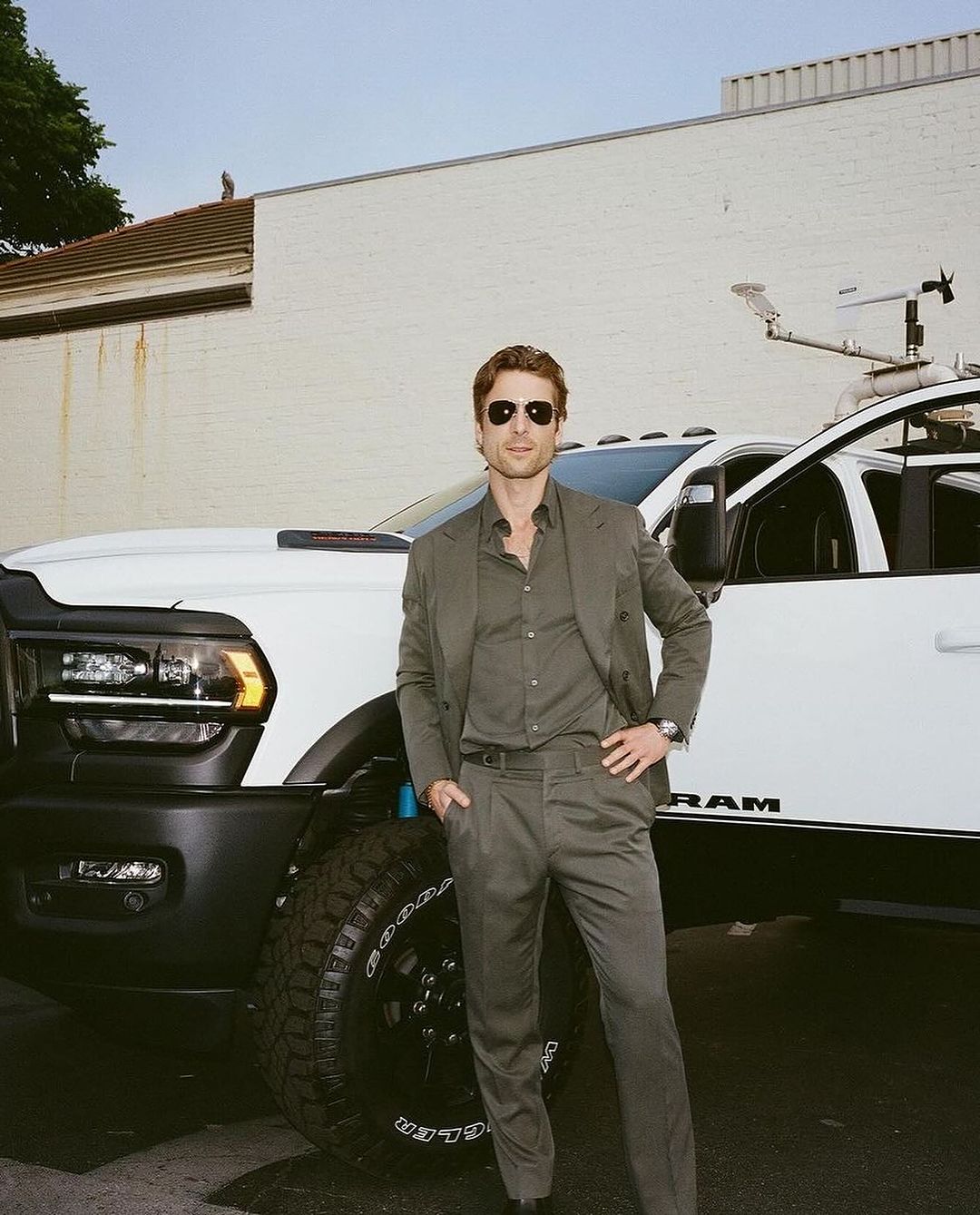 Adult male in sunglasses and gray suit poses confidently near a white RAM truck outdoors with a neutral building backdrop.