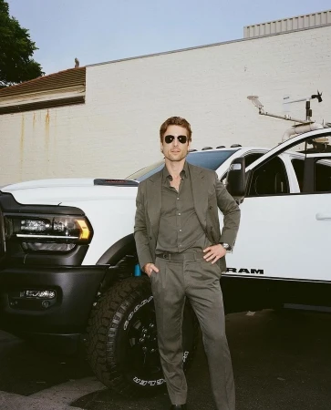 Adult male in sunglasses and gray suit poses confidently near a white RAM truck outdoors with a neutral building backdrop.