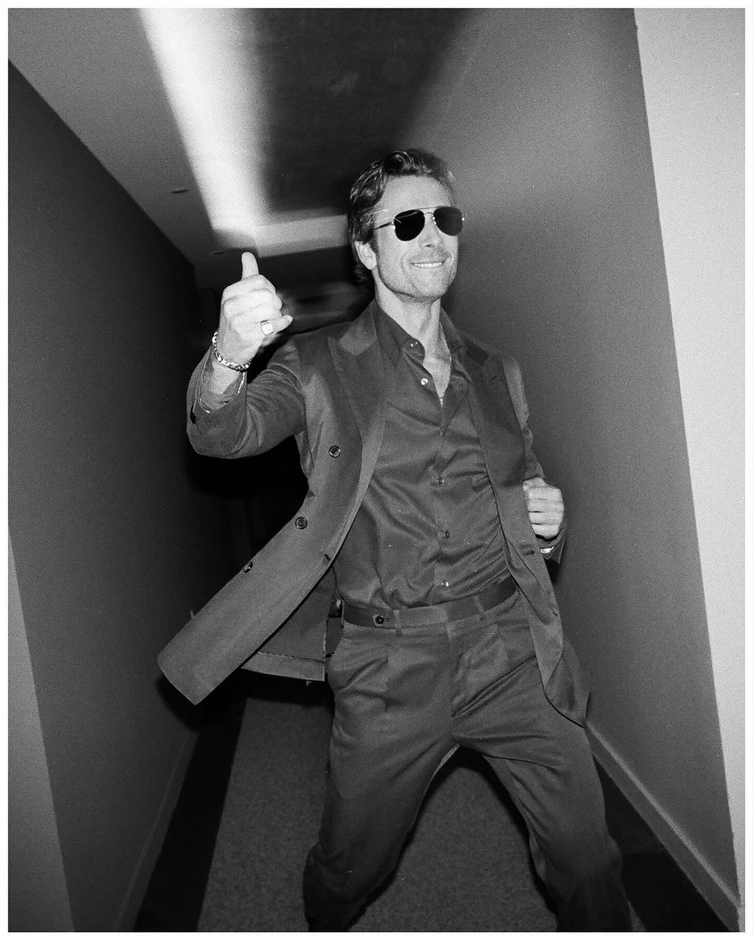 Black and white shot of a smiling man in a suit giving a thumbs up, illuminated by bright light in an indoor corridor.