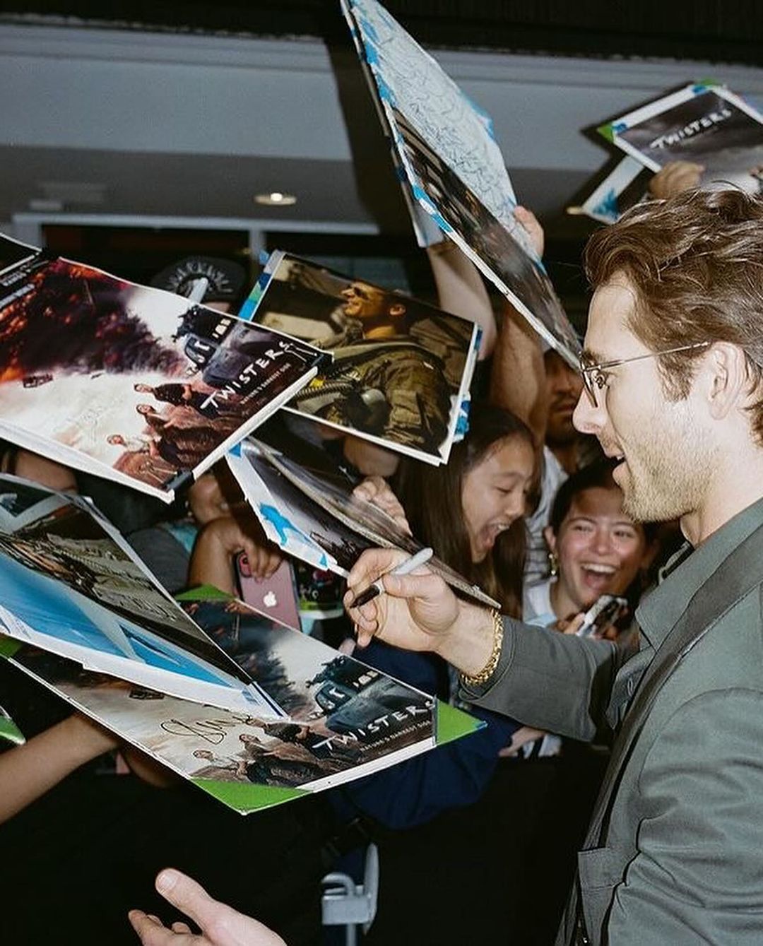 Actor Glen Powell signing autographs on 'Twisters' movie posters surrounded by fans at an event, smiling, wearing glasses.