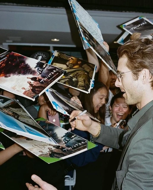 Actor Glen Powell signing autographs on 'Twisters' movie posters surrounded by fans at an event, smiling, wearing glasses.